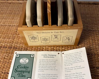 May include: A wooden caddy containing four rectangular ceramic cookie stamping tiles with embossed designs. A label on the front displays the text Tile Designs In This Set with illustrations of snowflakes and snowmen. An instruction sheet is included.