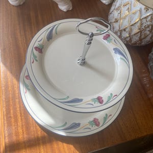 May include: A two-tiered serving stand made of cream-colored ceramic plates with a floral pattern featuring red flowers and blue leaves. The stand has a central silver-toned metal handle with a loop at the top for easy carrying.
