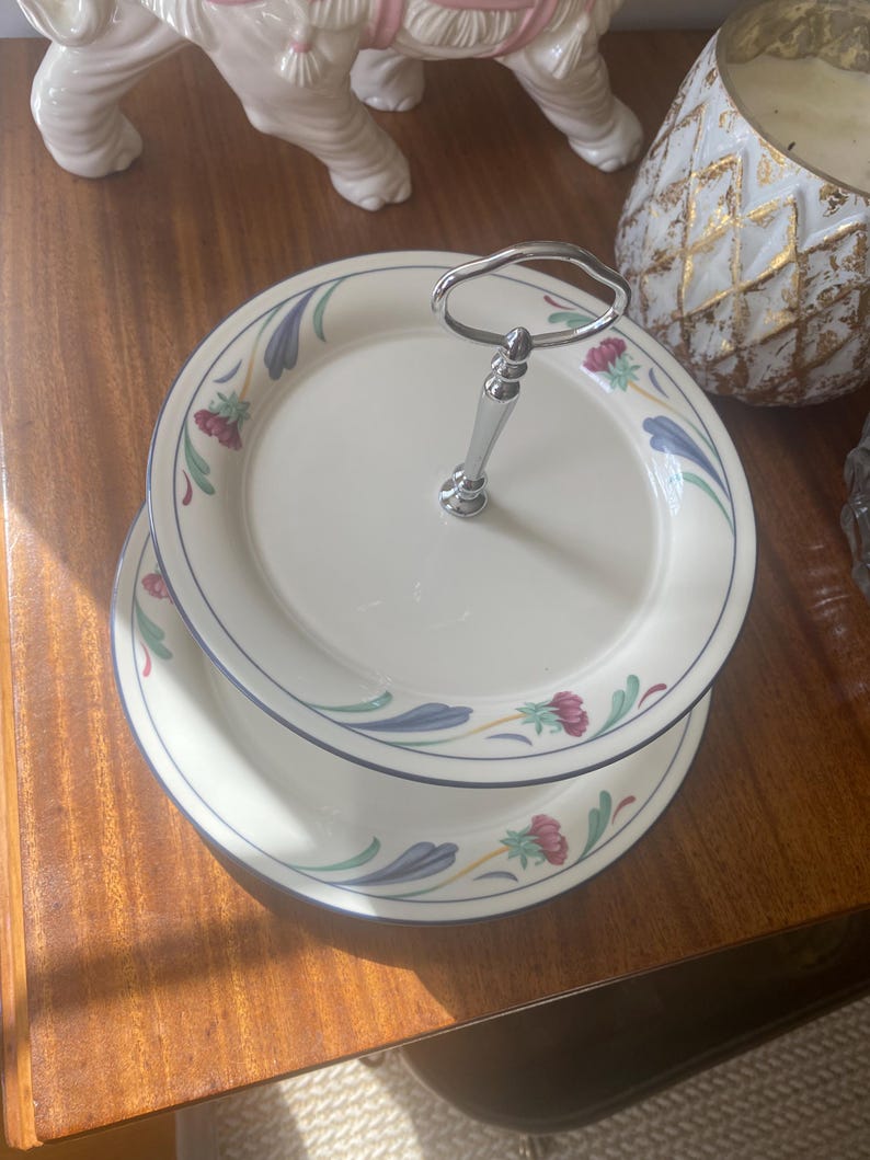 May include: A two-tiered serving stand made of cream-colored ceramic plates with a floral pattern featuring red flowers and blue leaves. The stand has a central silver-toned metal handle with a loop at the top for easy carrying.