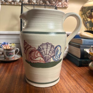 May include: A cream-colored ceramic pitcher featuring a hand-painted design of three seashells in shades of pink, purple, and blue. The pitcher has a curved handle, a pouring spout, and horizontal green bands encircling the top and bottom sections.