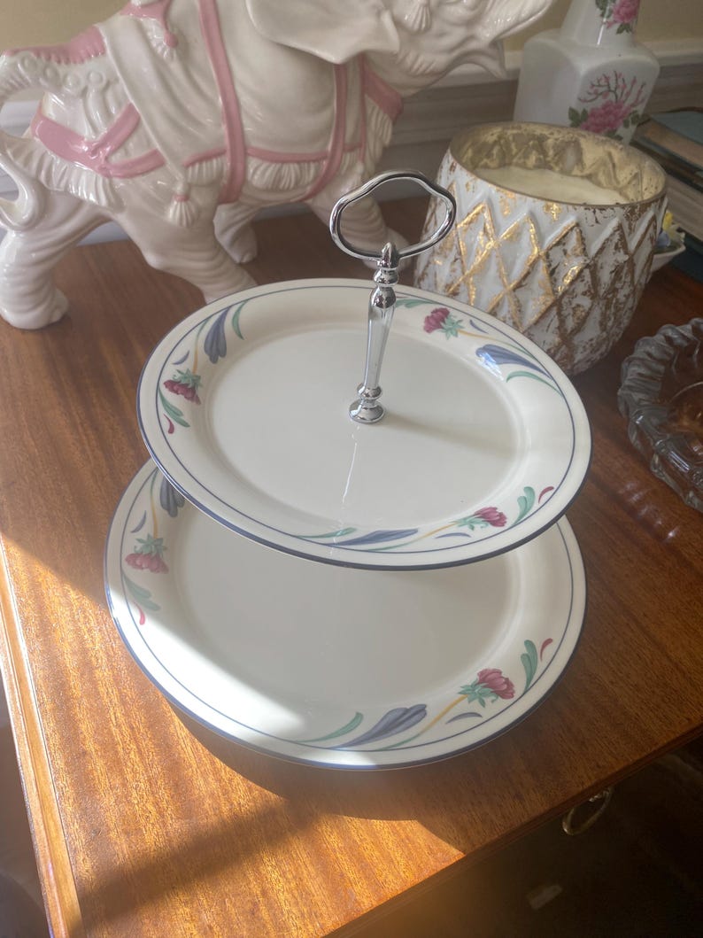 May include: A two-tiered serving stand featuring cream-colored ceramic plates with a delicate floral pattern in shades of blue, green, and pink. The tiers are connected by a central polished silver-tone metal handle with a loop at the top.