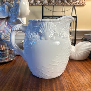 May include: A white ceramic pitcher featuring a raised relief design of seashells and a textured net pattern across the body. The pitcher has a curved handle and a pouring spout, with the shell motifs extending along the upper rim and handle.