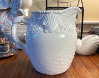 May include: A white ceramic pitcher featuring a raised relief design of seashells and a textured net pattern across the body. The pitcher has a curved handle and a pouring spout, with the shell motifs extending along the upper rim and handle.