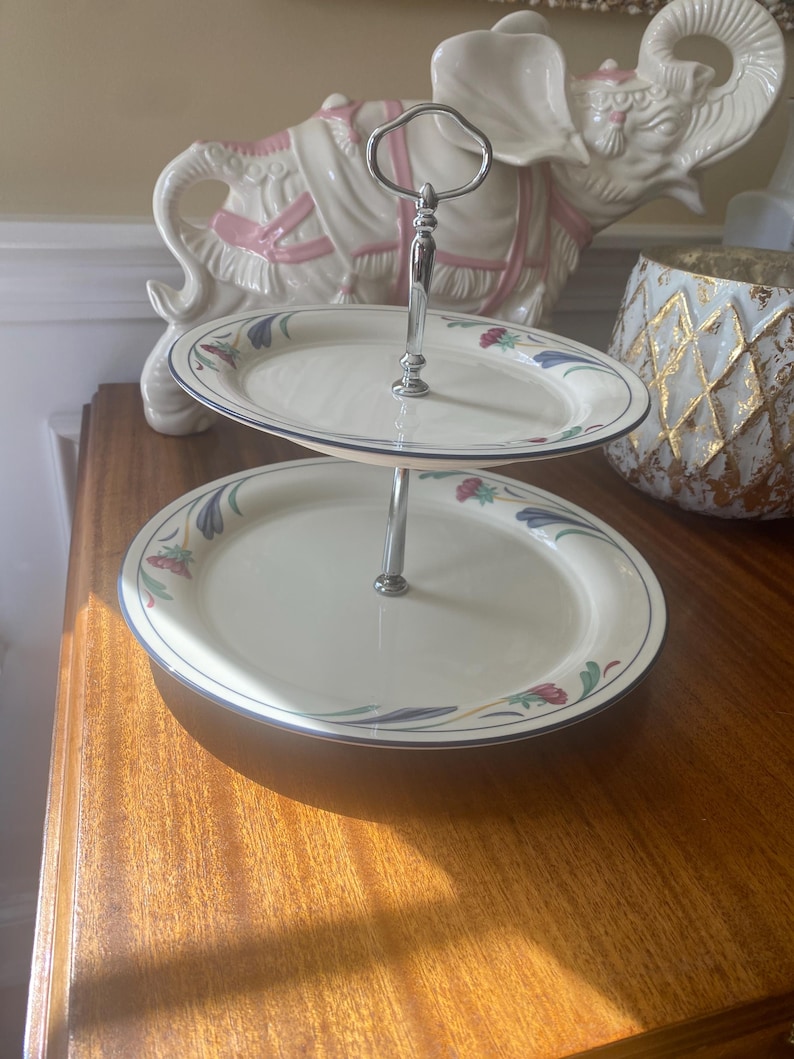 May include: A two-tiered serving stand featuring cream-colored ceramic plates with a floral pattern of blue, red, and green accents along the rims. The plates are connected by a central silver-toned metal rod with a looped handle at the top.