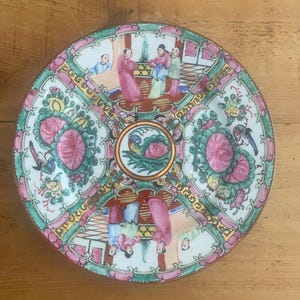 May include: A round, divided serving dish with a detailed, hand-painted design. The dish features a central medallion and four sections, each with a different scene of figures, flowers, and birds. The color palette includes pink, green, gold, and white.