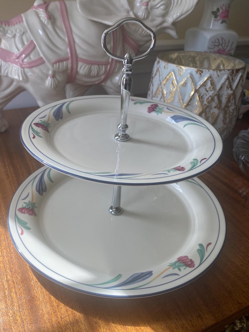 May include: Two-tiered serving tray with a silver handle. The white plates have a blue border and floral design in red, green, and blue. The tray is designed for serving desserts or appetizers.