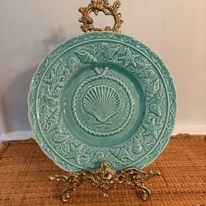 May include: A round, turquoise ceramic plate featuring a central embossed scallop shell surrounded by a rope border. The rim is decorated with raised starfish, various seashells, and coral motifs. The plate is displayed on an ornate gold metal stand.