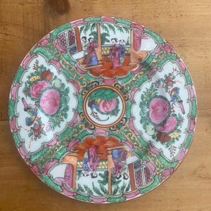 May include: A round, antique porcelain plate with a multi-sectioned design. The plate features a central floral motif and four surrounding sections with scenes of figures in traditional dress. The color palette includes pink, green, and gold accents.