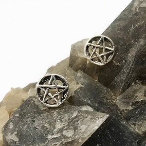 May include: A pair of silver stud earrings, each with a pentagram design. The earrings have a circular shape with an open star pattern. They are shown on a dark, textured surface.
