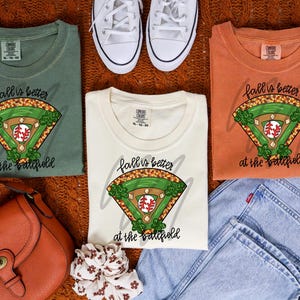 May include: Three t-shirts in green, cream, and orange, each with a baseball field graphic and the text "fall is better at the ballfield." The shirts are paired with jeans, white sneakers, and a brown purse, suggesting a casual, autumnal theme.