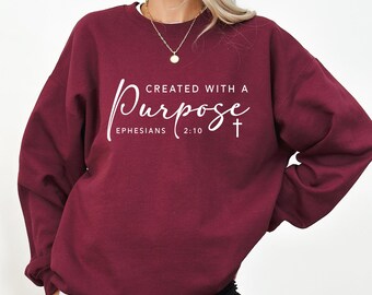 May include: A maroon sweatshirt with the text "CREATED WITH A Purpose" in white script. The text includes "Ephesians 2:10" and a cross symbol. The sweatshirt has a crew neck and long sleeves.