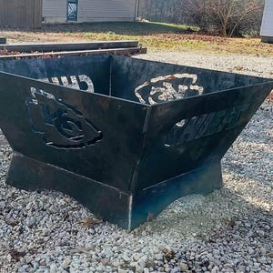 May include: A black metal fire pit with a square shape and angled sides. The sides feature cut-out designs of a team logo and the word "CHIEFS". The fire pit is sitting on a bed of small gray stones.