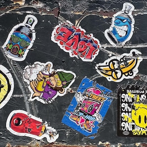 May include: A selection of vibrant graffiti stickers adhered to a dark, textured wall. The stickers depict spray paint cans, cartoon figures, and the word "LOVE" in red lettering. The stickers show signs of wear and tear, contributing to a street art style.