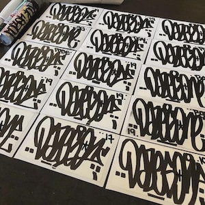 May include: A selection of black and white graffiti stickers, each displaying a unique stylized lettering design. The stickers are laid out on a surface, with spray paint cans partially visible. The graffiti style features bold lines and artistic flair.