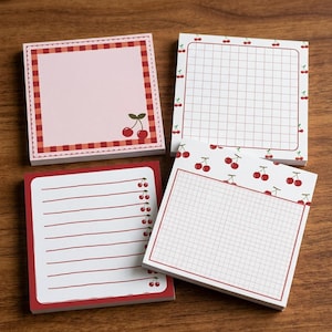 May include: Four square sticky note pads featuring cherry motifs. One pad has a pink center with a red checkered border, one has a white grid with cherry borders, one has red lined paper with cherries, and one has a white grid with cherry patterns.