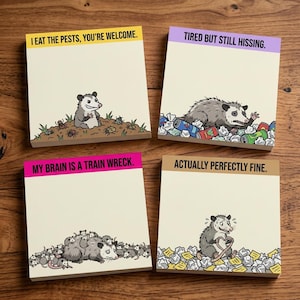 May include: Four square sticky note pads featuring opossum illustrations and text. Top left: yellow header, 'I EAT THE PESTS, YOU'RE WELCOME.' Top right: purple header, 'TIRED BUT STILL HISSING.' Bottom left: pink header, 'MY BRAIN IS A TRAIN WRECK.' Bottom right: tan header, 'ACTUALLY PERFECTLY FINE.'