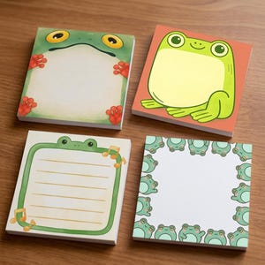 Puede incluir: Cuatro blocs de notas adhesivas cuadrados con ilustraciones de ranas. Un bloc muestra una cara de rana con pies rojos, otro una rana de dibujos sobre naranja, uno rayado con notas musicales y otro con un borde de pequeñas ranas verdes.