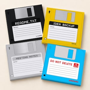 Puede incluir: Cuatro posavasos cuadrados diseñados como disquetes antiguos en negro, amarillo, gris y azul. Cada uno tiene una etiqueta blanca con texto impreso que incluye README.TXT, 1.44MB, IDEA BACKUP, MEETING NOTES y DO NOT DELETE con un icono de advertencia.