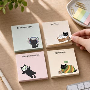 May include: Four square sticky note pads featuring black cat illustrations. One light green pad says In my own world with a cat wearing headphones. A beige pad says Me Time with a cat in a bathtub. A pink pad says Self-care in progress with a cat wearing a face mask. A cream pad says Recharging with a cat wrapped in a blanket.