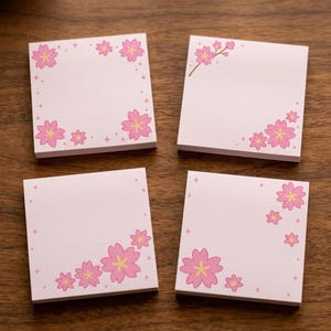 Puede incluir: Cuatro notas adhesivas cuadradas de color rosa claro dispuestas en cuadrícula. Cada nota presenta un patrón único de flores de cerezo rosas con centros amarillos y pequeños detalles de estrellas de cuatro puntas sobre el papel.