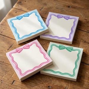 Puede incluir: Cuatro blocs de notas adhesivas cuadrados con centros de color crema. Cada bloc presenta un borde decorativo en un color pastel diferente: azul, morado, rosa y verde. Un lazo a juego está ilustrado en la parte superior central de cada marco ondulado.