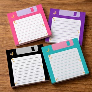 May include: Four square sticky note pads designed to resemble floppy disks in pink, purple, black, and teal. Each pad features a white lined writing area, a top label section, and small yellow crescent moon and star icons printed on the corners.