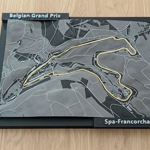 May include: A 3D map of the Spa-Francorchamps race track, home of the Belgian Grand Prix. The track is highlighted in gold against a gray topographical background. The map is set in a black frame with the text "Belgian Grand Prix" and "Spa-Francorchamps".