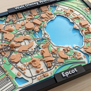 Può includere: Una mappa stampata in 3D di Walt Disney World, che mostra la planimetria del parco e le sue attrazioni. La mappa è incorniciata in nero, con i nomi "Walt Disney World" ed "Epcot" visualizzati. La mappa include un lago blu e aree verdi.