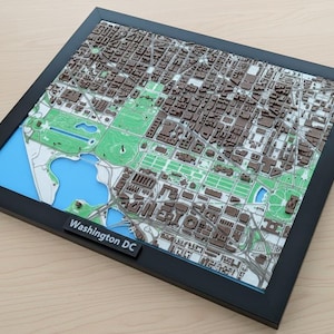 May include: A framed 3D map of Washington, D.C. The map features a detailed relief of the city's buildings, parks, and waterways, with brown, green, and blue color accents. The frame is black, and the text "Washington DC" is visible.