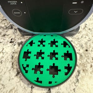 May include: A green and black round puzzle-shaped drip tray for a coffee maker. The tray has a textured surface and is designed to catch drips from the coffee maker.