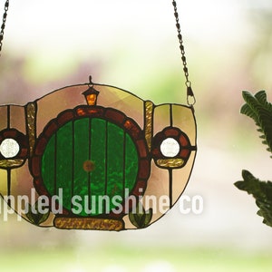 May include: A stained glass window hanging with a chain, depicting a green door with a round handle, surrounded by brown brick and a yellow light above. The text "dappled sunshine co" is visible at the bottom.