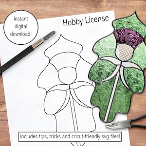May include: A black and white line drawing of a thistle flower with a green and purple stained glass thistle flower next to it. The text "Hobby License" is at the top of the page. The text "includes tips, tricks and cricut-friendly svg files!" is at the bottom of the page.