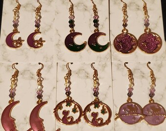 May include: Six pairs of dangle earrings with a moon and star theme. The earrings feature a variety of colors, including purple, green, and gold. The earrings are made with metal and glass beads.