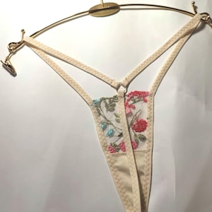 May include: A white lace thong with floral embroidery in pink, blue and green. The thong is hanging on a gold hanger.