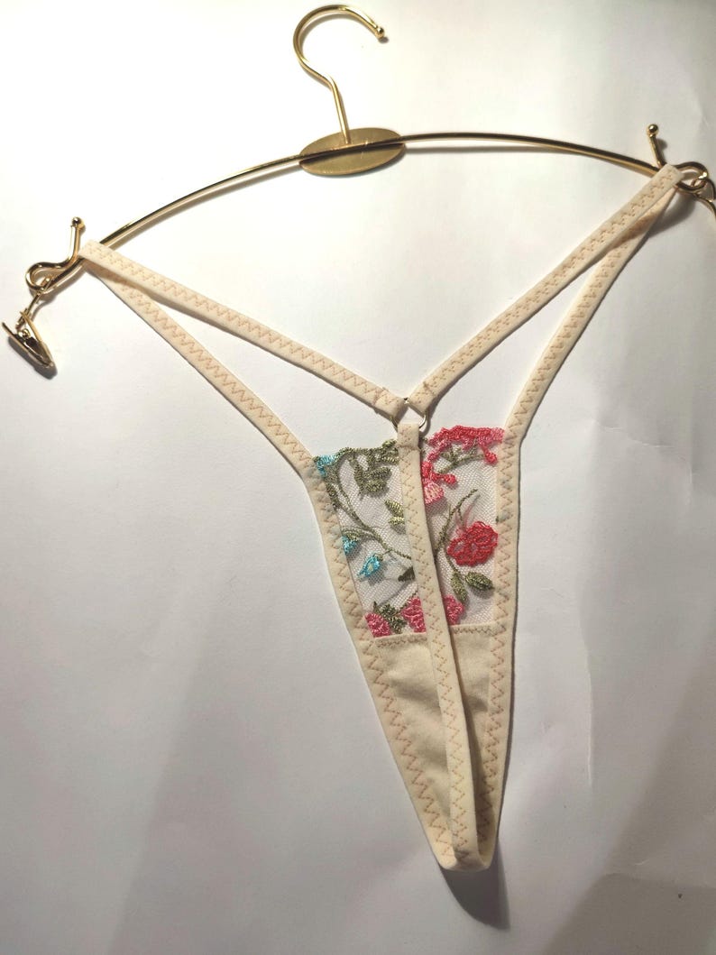 May include: A white lace thong with floral embroidery in pink, blue and green. The thong is hanging on a gold hanger.
