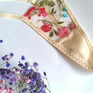 May include: A delicate beige lingerie with floral embroidery in shades of pink, blue, and green. The embroidery features a floral pattern with leaves and flowers.