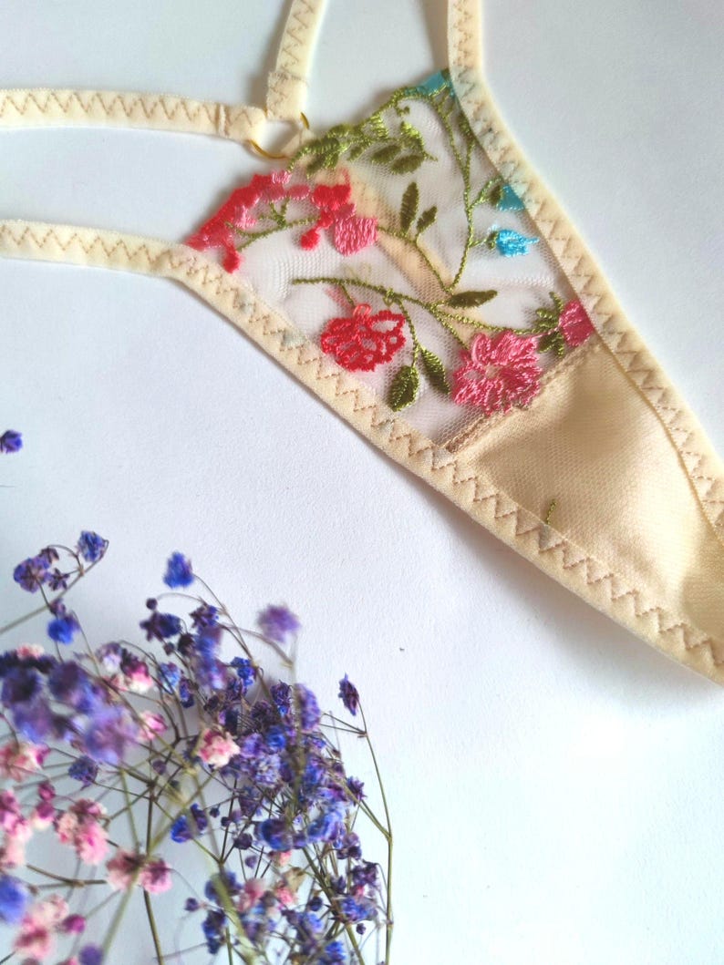 May include: A delicate beige lingerie with floral embroidery in shades of pink, blue, and green. The embroidery features a floral pattern with leaves and flowers.