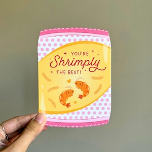 May include: A greeting card with a pink and white polka dot border and a yellow center. The card features two cartoon shrimp and the text "You're Shrimply the Best!"