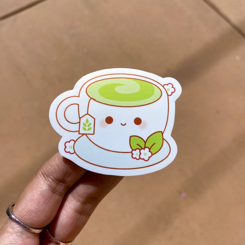 Pode incluir: Um adesivo branco com uma ch&aacute;vena de ch&aacute; de desenho animado cheia de ch&aacute; verde. A ch&aacute;vena tem um rosto sorridente, um saquinho de ch&aacute; e &eacute; decorada com flores. O adesivo est&aacute; numa superf&iacute;cie castanha.