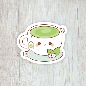 Pode incluir: Um autocolante branco com uma ch&aacute;vena de ch&aacute; de desenho animado sorridente, cheia de ch&aacute; verde, com um saquinho de ch&aacute;, flores e folhas. O autocolante est&aacute; sobre um fundo claro com gr&atilde;o de madeira.