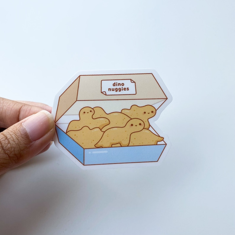 Peut inclure: Un autocollant repr&eacute;sentant une bo&icirc;te de nuggets de poulet en forme de dinosaures de dessin anim&eacute;. La bo&icirc;te est marron clair avec une base bleue et les mots "dino nuggies" sur une petite &eacute;tiquette. Les p&eacute;pites sont marron clair avec des visages souriants.