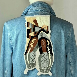 Puede incluir: Chaqueta vaquera azul claro con un parche bordado rectangular grande en la espalda. El parche presenta un diseño abstracto en tonos marrón, gris, negro y crema, con la palabra "Ballet" en cursiva. La chaqueta tiene cuello clásico y botones.