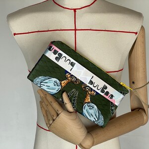 Könnte beinhalten: Eine grüne Clutch mit Rautenmuster und gelbem Reißverschluss. Die Clutch zeigt eine sich wiederholende Illustration einer vogelartigen Kreatur und den Text "buuBoo" auf einem weißen Band. Zwei kleine Federn sind am Reißverschluss befestigt.