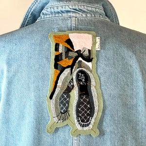 May include: A light blue denim jacket with an embroidered patch. The patch features a design with black, grey, yellow, and pink. The patch is in the shape of a shoe sole and has the word "Ballet" on it.