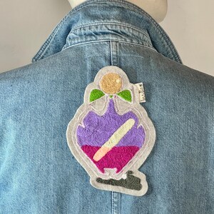Peut inclure: Un écusson brodé en forme de flacon de potion, orné de couleurs violettes, roses et jaunes. Il est fixé sur une veste en jean bleu clair, mettant en valeur le design décoratif.