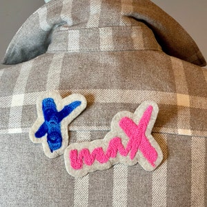 Op de afbeelding: Een grijs geruit flanellen overhemd met twee viltpatches die op de achterkant zijn genaaid. De patches hebben de vorm van de woorden "Dank je" met de letters "D" en "K" in blauw en roze garen.