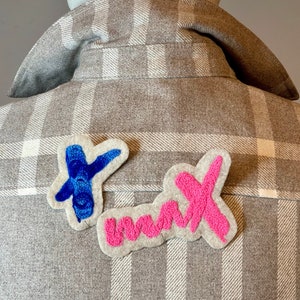 Op de afbeelding: Een grijs geruit flanellen overhemd met twee viltpatches die op de achterkant zijn genaaid. De patches hebben de vorm van de woorden "Dank je" met de letters "D" en "K" in blauw en roze garen.