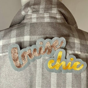 Può includere: Una camicia di flanella a quadri grigi e bianchi con due toppe ricamate. Le toppe hanno la forma di parole. La toppa superiore dice "cruise" in filo marrone e beige. La toppa inferiore dice "chic" in filo giallo.