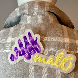 Peut inclure: Une chemise en flanelle à carreaux bruns et blancs avec deux patchs brodés. Le patch du haut est violet et dit "right", le patch du bas est jaune et dit "now".