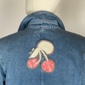 Peut inclure: Une veste en jean bleue avec un patch brodé de cerises rose et blanc au dos.
